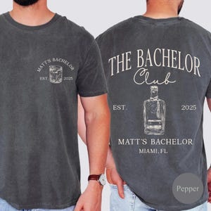 May include: Dark gray t-shirt with a front graphic of a cocktail and the text "MATT'S BACHELOR EST. 2025". The back of the shirt features a bottle graphic and the text "THE BACHELOR CLUB EST. 2025 MATT'S BACHELOR MIAMI, FL".