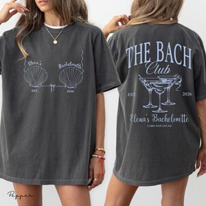 May include: Dark gray t-shirt with light blue graphics. The front features seashell designs and text: "Elena's Bachelorette EST. 2026." The back displays "THE BACH Club" and martini glasses, with "Elena's Bachelorette CABO SAN LUCAS EST. 2026."