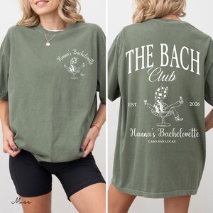 May include: Moss green t-shirt with white text and graphic. The front features "Hanna's Bachelorette" and a graphic of a person in a chair. The back says "THE BACH CLUB" with "EST. 2026" and "Hanna's Bachelorette CABO SAN LUCAS".