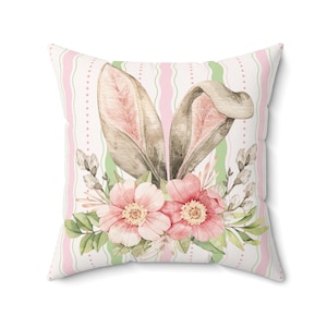 May include: Square throw pillow with a watercolor design featuring bunny ears, pink flowers, and green leaves. The background has pink and green vertical stripes. A decorative accent for Easter or spring.