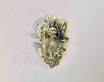 May include: A gold enamel pin depicting a skeleton in a coffin, framed by floral motifs. The pin's design includes black and white detailing, contrasting with the gold. This decorative pin is a unique accessory, suitable for adding a touch of gothic flair.