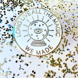 May include: White enamel pin with gold lettering and outline. The pin features a crystal ball with an eye inside and the text "Long Live All The Magic We Made".