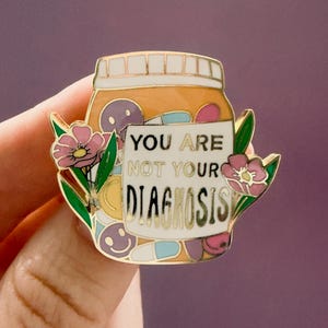 May include: A gold-toned metal enamel pin shaped like a medicine bottle filled with colorful pills and smiley faces. The white label on the front features the black text "YOU ARE NOT YOUR DIAGNOSIS." Pink flowers with green leaves flank the bottle.