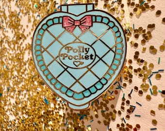 Puede incluir: Un compacto Polly Pocket en forma de corazón azul y dorado con un lazo rosa y un patrón de cuadrícula blanca. El compacto tiene las palabras "Polly Pocket" escritas en él.