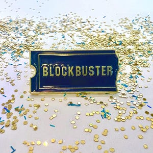 May include: A gold-coloured pin with a blue background and the word "BLOCKBUSTER" in gold letters. The pin is on a white surface with gold and blue glitter.