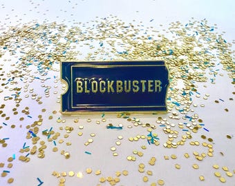 May include: A gold-colored pin with a blue background and the word "BLOCKBUSTER" in gold letters. The pin is on a white surface with gold and blue glitter.