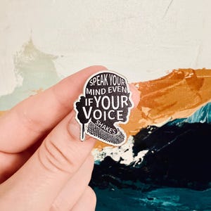 May include: A silver-colored enamel pin with a black and white design. The pin features a silhouette of a person's head and shoulders with the text "Speak Your Mind Even If Your Voice Shakes".