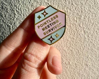May include: A shield-shaped enamel pin with a gold border. The pin features a light pink and blue color scheme with the words "POINTLESS MEETING SURVIVOR" in gold lettering. Decorative gold stars and shapes are also present.