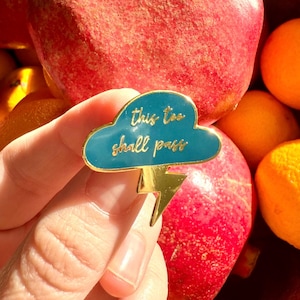 May include: A gold-colored enamel pin with a blue cloud and a lightning bolt. The cloud has the text "this too shall pass" written on it.