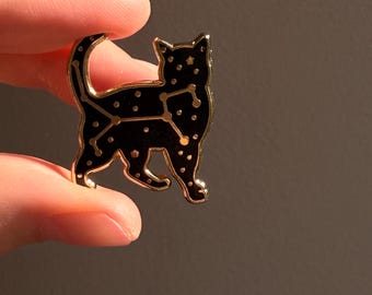 May include: A gold-coloured metal cat-shaped pin with a black enamel fill. The pin features a constellation design with small white dots. The pin is held between two fingers against a dark background.