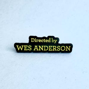 Puede incluir: Pin de esmalte negro y amarillo con el texto "Directed by WES ANDERSON".