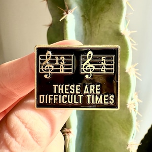 Könnte beinhalten: Schwarze Emaille-Anstecknadel mit goldenen Akzenten, die Noten und den Text "These are difficult times" zeigen.