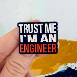 May include: A rectangular enamel pin with a black background and white text that reads TRUST ME I'M AN. The word ENGINEER is printed in bold orange capital letters at the bottom of the pin. The pin has a black border and a smooth finish.