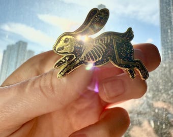 Op de afbeelding: Gouden metalen pin van een konijnenskelet met glitteraccenten. Het konijn rent en heeft een ster op zijn oor.