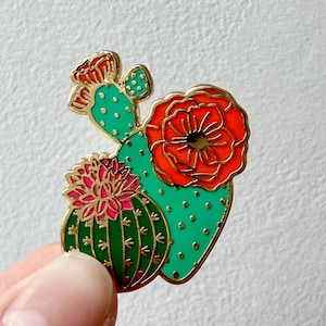 May include: A gold-tone enamel pin featuring a green cactus with a red flower and a smaller pink cactus with green spines. The cactus is outlined in gold.