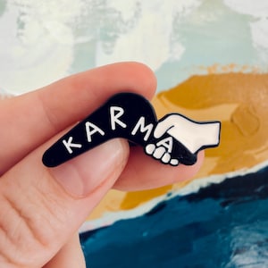 May include: A black enamel pin shaped like a boomerang with the white text KARMA printed on it. The right end of the pin features a white hand reaching out to grasp a black hand, symbolizing a handshake or connection in a high-contrast design.