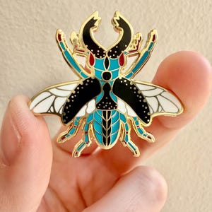 Puede incluir: Pin de escarabajo dorado con esmalte negro, blanco y turquesa. El escarabajo tiene alas y un diseño detallado.