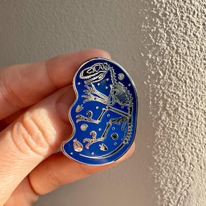 May include: A blue enamel pin featuring a silver dinosaur skeleton with small white bones and a blue background. The dinosaur is in a fetal position with its head tilted back.