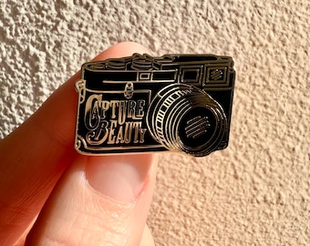 Puede incluir: Un pin de esmalte negro y dorado con forma de cámara vintage. El pin presenta las palabras "CAPTURE BEAUTY" en una fuente elegante. La lente y los detalles de la cámara están resaltados en dorado.