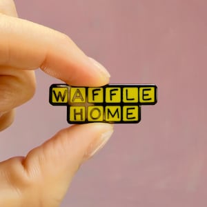 Peut inclure: Une broche en émail rectangulaire présentant le texte WAFFLE HOME en lettres noires sur un fond jaune. Le mot WAFFLE est sur la ligne supérieure et le mot HOME sur la ligne inférieure, chaque lettre étant dans un carré jaune à bordure noire.