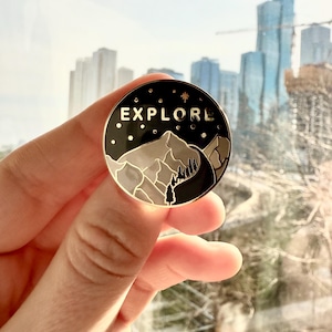 May include: A gold-toned enamel pin with a black background featuring a mountain range silhouette, a pine tree, and the word "EXPLORE" in white lettering. The pin has a star and small white dots around the edge.