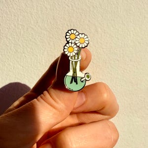 Può includere: Uno spillo in smalto argentato con un bong di vetro verde con un mazzo di margherite bianche al suo interno. Il bong ha un bocchino verde e uno stelo bianco.