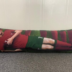 May include: A long rectangular pillow with a green and red striped background. A soccer player is lying on the pillow wearing a red jersey with the number 7 on it. The player is wearing green shorts and red socks.