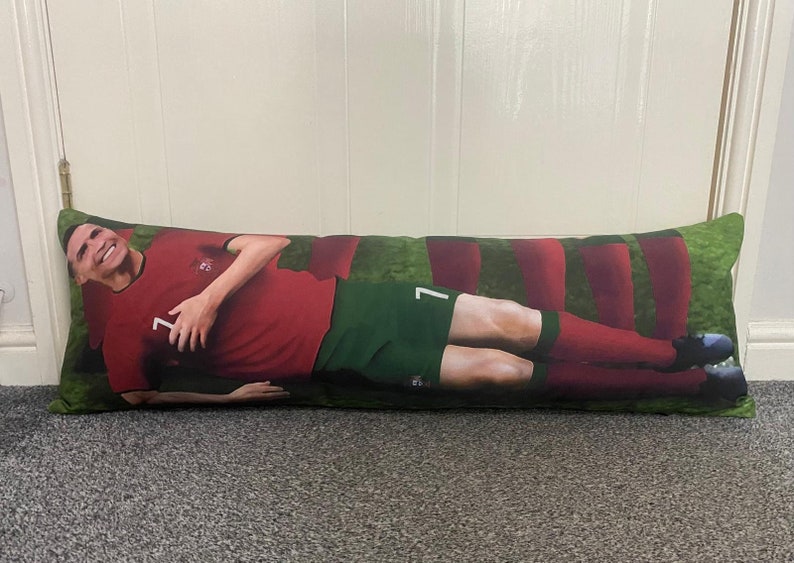 May include: A long rectangular pillow with a green and red striped background. A soccer player is lying on the pillow wearing a red jersey with the number 7 on it. The player is wearing green shorts and red socks.