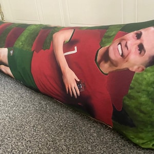 May include: A long red and green pillow with a smiling man's face on it. The man is wearing a red soccer jersey with a number 7 on it and the Portuguese flag on the chest. The pillow is made of soft fabric and is perfect for lounging.