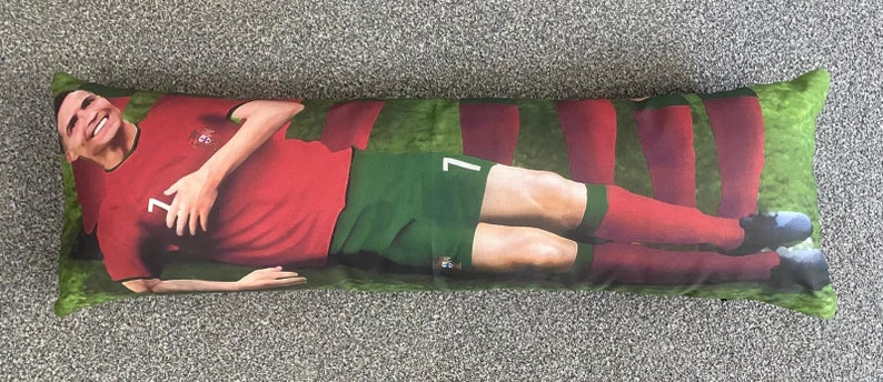 May include: A long body pillow featuring a soccer player in a red and green jersey with the number 7 on it. The player is lying on his back with his arms outstretched and legs crossed. The pillow is green with red stripes.