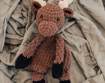 May include: A handmade crochet moose plush toy. The moose is brown with black hooves and cream-colored antlers. The toy is lying on a gray, textured blanket. The moose has a friendly expression.