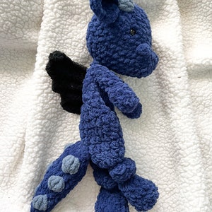 May include: A blue crocheted dragon plush toy with black wings and gray accents. The dragon is lying on a white fluffy blanket.