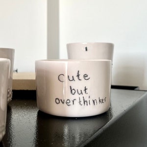 May include: A small, cylindrical, off-white ceramic mug featuring the hand-lettered black text "Cute but overthinker" on the front. The mug has a smooth, glossy finish and a simple, straight-sided design with a flat base and an open top.