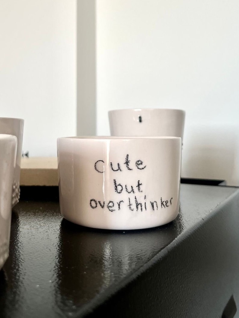 May include: A small, cylindrical, off-white ceramic mug featuring the hand-lettered black text "Cute but overthinker" on the front. The mug has a smooth, glossy finish and a simple, straight-sided design with a flat base and an open top.