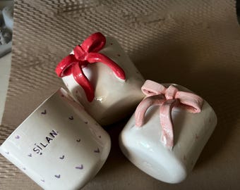 May include: Three white ceramic mugs with a heart pattern and a bow design. One mug has the name "ŞILAN" printed on it. The bows are red and pink. The mugs are on a brown surface.
