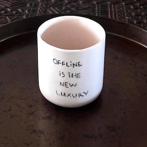 May include: A white ceramic cup with a smooth, matte finish and a cylindrical shape. The front of the cup features the black handwritten text "OFFLINE IS THE NEW LUXURY" in a simple, sans-serif style. The interior of the cup is also white.