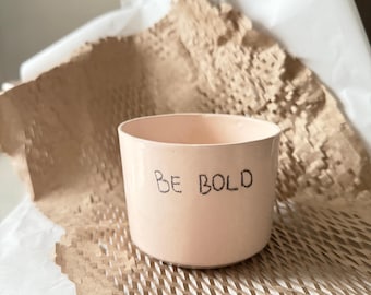 May include: A pink ceramic cup with the words "BE BOLD" written in black marker.