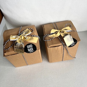 May include: Two square cardboard boxes in a light brown color, each tied with black and white baker's twine and topped with a metallic gold ribbon. A circular black sticker on the front of each box features the white text HAND MADE WITH LOVE.