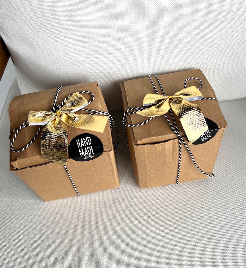 May include: Two square cardboard boxes in a light brown color, each tied with black and white baker's twine and topped with a metallic gold ribbon. A circular black sticker on the front of each box features the white text HAND MADE WITH LOVE.
