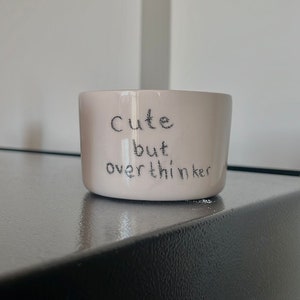May include: A light pink ceramic mug with a smooth, glossy finish. The text "Cute but overthinker" is printed on the front in a dark, handwritten-style font. The mug has a simple, cylindrical shape with a flat base and no visible handle.