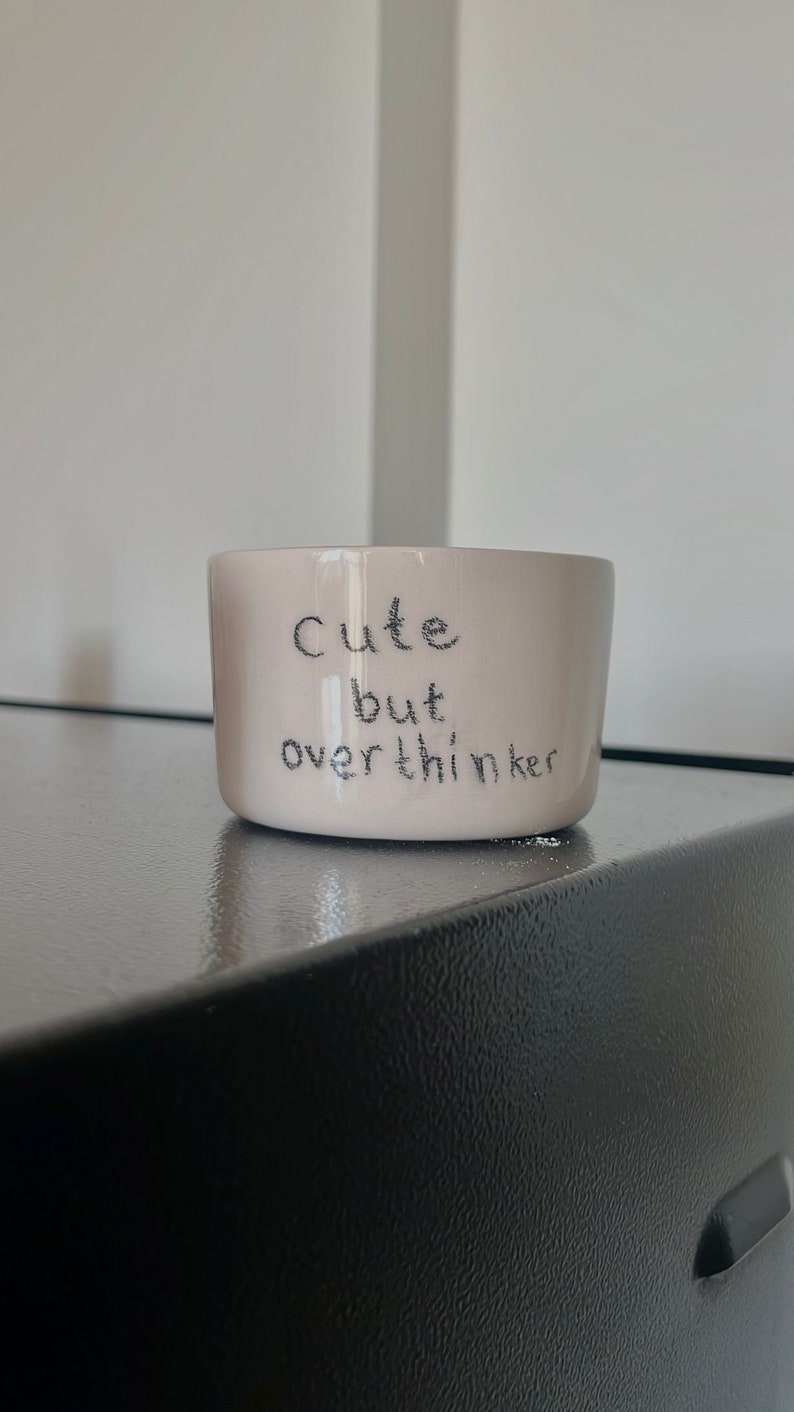 May include: A light pink ceramic mug with a smooth, glossy finish. The text "Cute but overthinker" is printed on the front in a dark, handwritten-style font. The mug has a simple, cylindrical shape with a flat base and no visible handle.