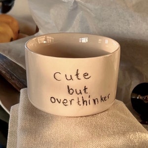 May include: A cream-colored ceramic mug with a smooth, cylindrical shape. The front of the mug features the black, hand-lettered text "Cute but overthinker" written in a casual, slightly textured font style across three lines.
