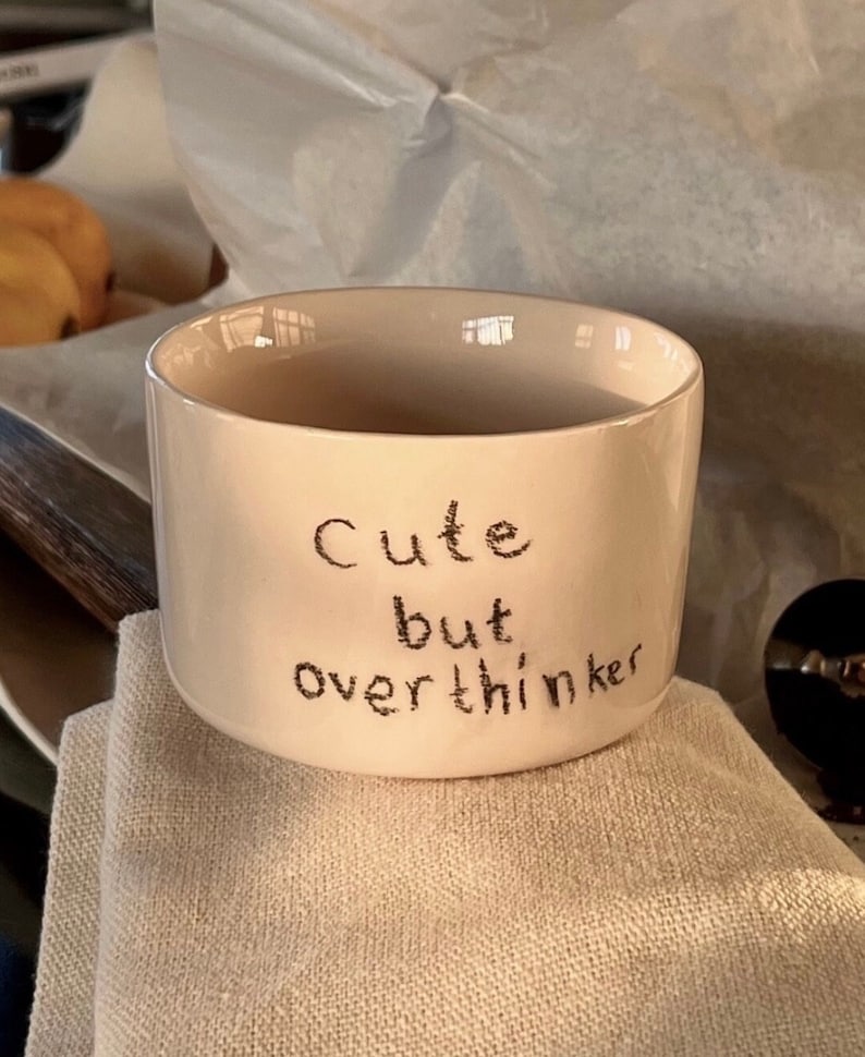 May include: A cream-colored ceramic mug with a smooth, cylindrical shape. The front of the mug features the black, hand-lettered text "Cute but overthinker" written in a casual, slightly textured font style across three lines.