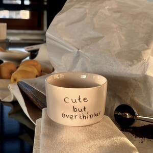 May include: A cream-colored ceramic mug with a straight-sided cylindrical shape. The exterior features the black handwritten text "Cute but overthinker" centered on the front. The mug has a smooth finish and a wide, open top rim.