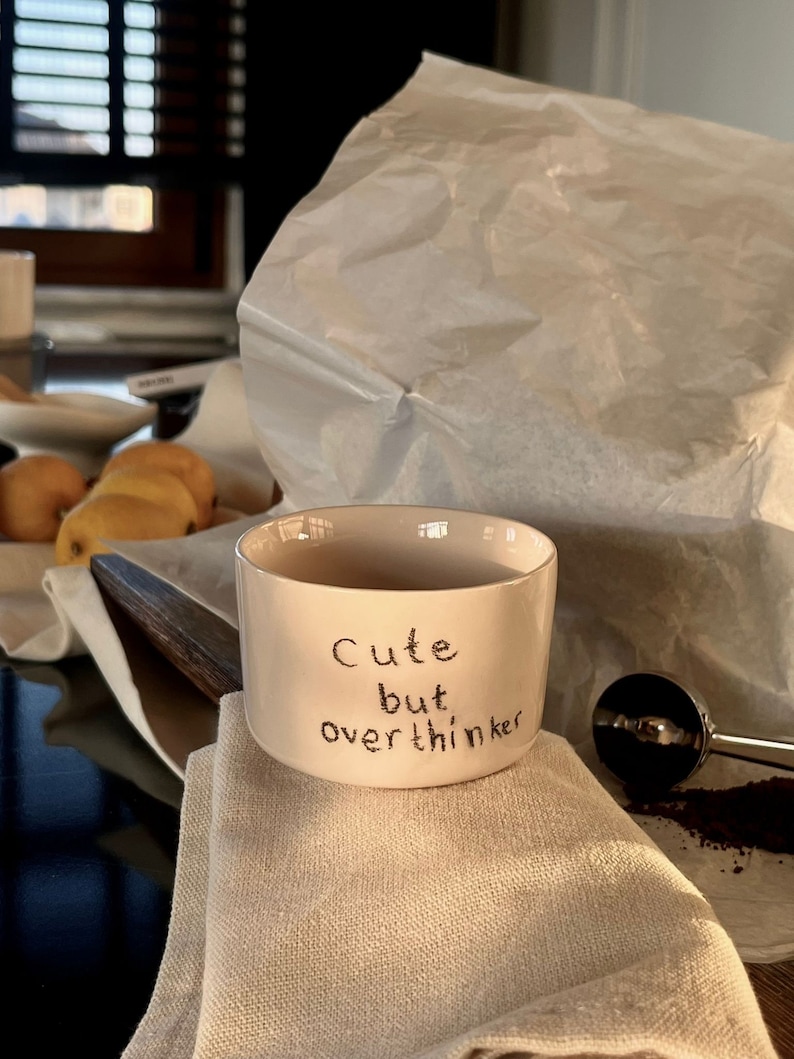 May include: A cream-colored ceramic mug with a straight-sided cylindrical shape. The exterior features the black handwritten text "Cute but overthinker" centered on the front. The mug has a smooth finish and a wide, open top rim.