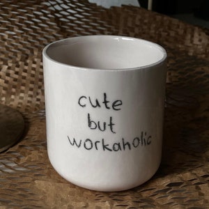 May include: White ceramic mug with black handwritten text that reads "cute but workaholic".