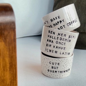 May include: Stacked white ceramic coffee mugs with black speckles and text. The mugs read "DON'T MAKE ME HAPPY NOT. COFFEE", "BEN HER ŞEYİ HALLEDERİM AMA ÖNCE BİR KAHVE İÇMEM LAZIM", and "CUTE BUT OVERTHINKER".
