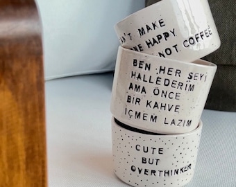 May include: Stacked white ceramic coffee mugs with black speckles and text. The mugs read "DON'T MAKE ME HAPPY NOT. COFFEE", "BEN HER ŞEYİ HALLEDERİM AMA ÖNCE BİR KAHVE İÇMEM LAZIM", and "CUTE BUT OVERTHINKER".
