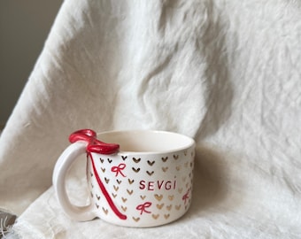 May include: A white ceramic mug with a gold heart pattern and the word "SEVGI" in brown. The mug features a red ribbon design and a red bow on the handle. The mug is on a white cloth.