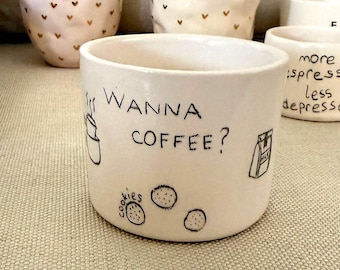 Könnte beinhalten: Eine Sammlung handgefertigter Keramik-Kaffeetassen. Eine Tasse zeigt den Text "WANNA COFFEE?" mit Illustrationen einer Kaffeetasse, Keksen und einem Milchkarton. Andere Tassen haben Herz- und Initialen-Designs.